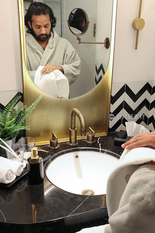 镜子里的印度男人穿着浴袍站在浴室的水槽前，用白色毛巾擦手，大理石浴室柜台上的金属盆里放着人造蕨类植物，浴室水槽，玻璃牙刷，法兰绒