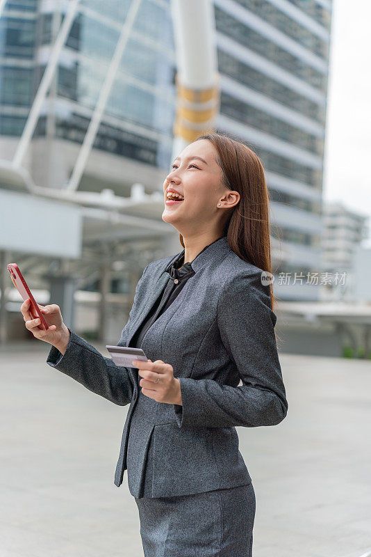 一名年轻的亚洲女商人穿着灰色的办公套装，在商业区用她的智能手机使用信用卡进行在线交易，她微笑着自信