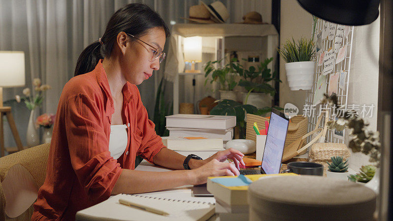 年轻的亚洲女性晚上穿着便服坐在书桌前，用电脑笔记本电脑在家里的客厅里思考创造工作的想法。居家办公