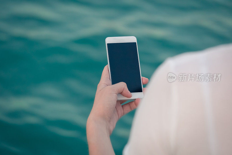 女性拿着空白屏幕的智能手机对着蓝色的海洋水面