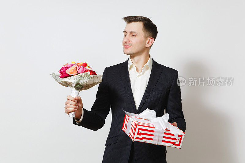 年轻英俊的男子在西装捧着一束美丽的玫瑰花，红礼盒与礼物隔离在白色的背景。情人节、国际妇女节生日节日概念。