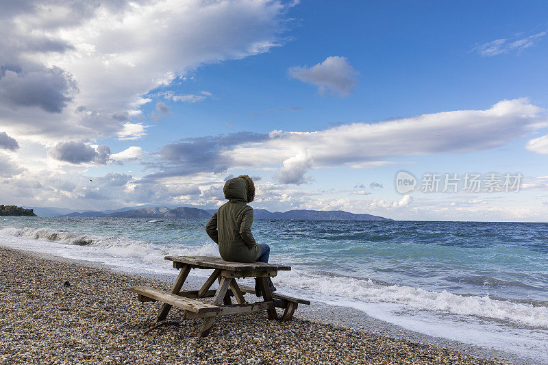 年轻女孩坐在海滩上的野餐桌上看着波涛汹涌的大海，波涛汹涌的大海，大风的天气