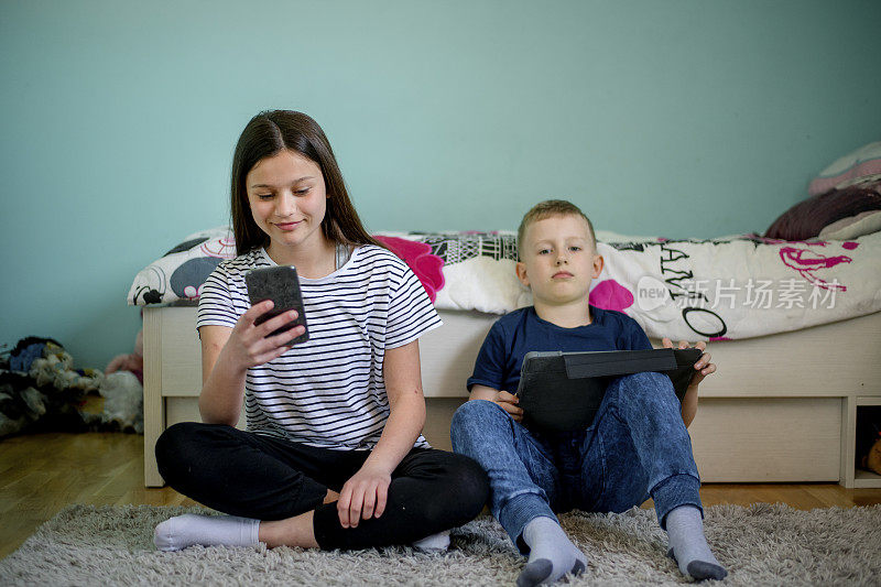 两个孩子拿着平板电脑和手机坐在家里的地板上