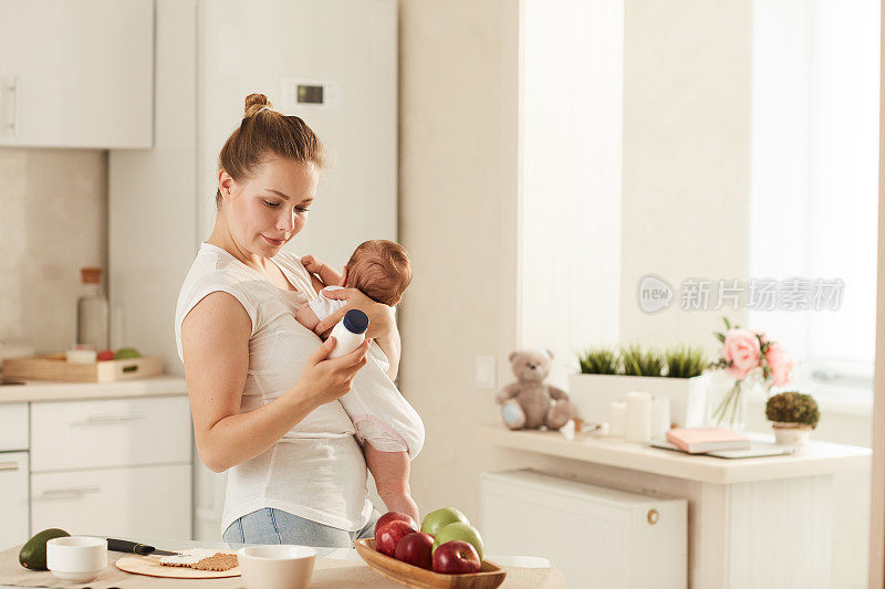 年轻的母亲站在厨房里，怀里抱着婴儿，阅读有关婴儿配方奶粉的资料