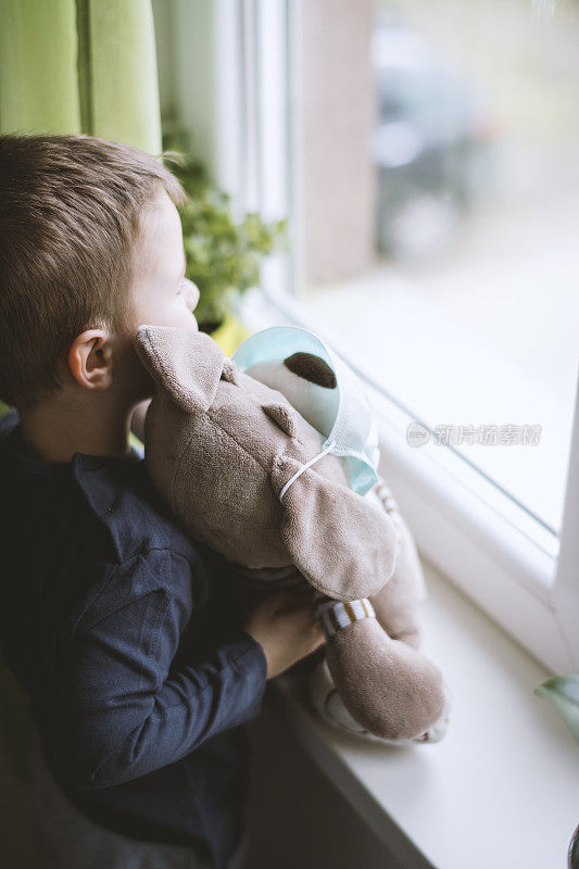 小男孩戴着防护口罩，带着闷热的玩具，从窗户往外看。预防传染病