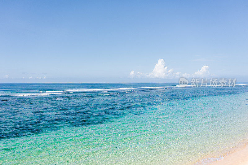 巴厘岛海滩的场景-蓝绿色的印度洋，白色的沙滩和蓝色的多云的天空在背景