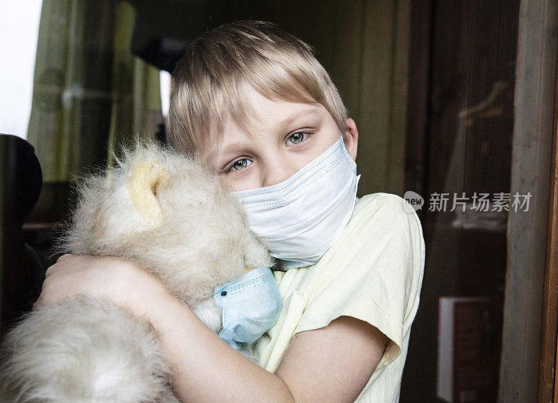 一个男孩带着他生病的泰迪熊，戴着医用防护口罩从窗户往外看。预防流行病。