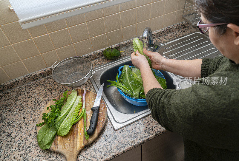 一个成熟的女人在家里的厨房水槽里清洗有机长叶莴苣，准备晚餐沙拉