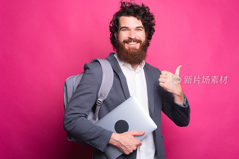 微笑的年轻人的肖像背包和手提电脑和显示拇指向上