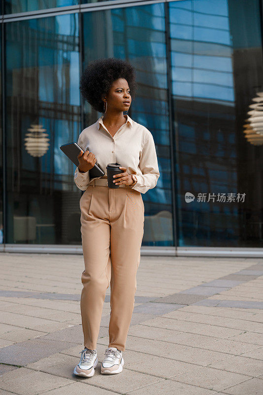 一位深色皮肤的年轻女子，身穿米色套装，端着一杯咖啡，拿着一台数码平板电脑，走在商业区的广场上，目光移开