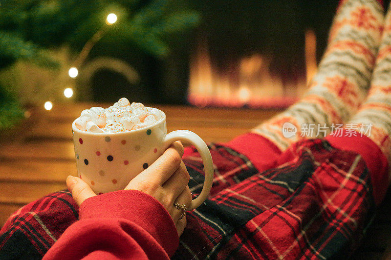 近距离照片中，一名无法辨认的女子穿着红色格子睡衣，穿着圣诞主题的图案袜子，脚踩满斑点的咖啡桌，杯子里是热巧克力，上面放着鲜奶油和迷你棉花糖，背景是一排瓦斯火焰