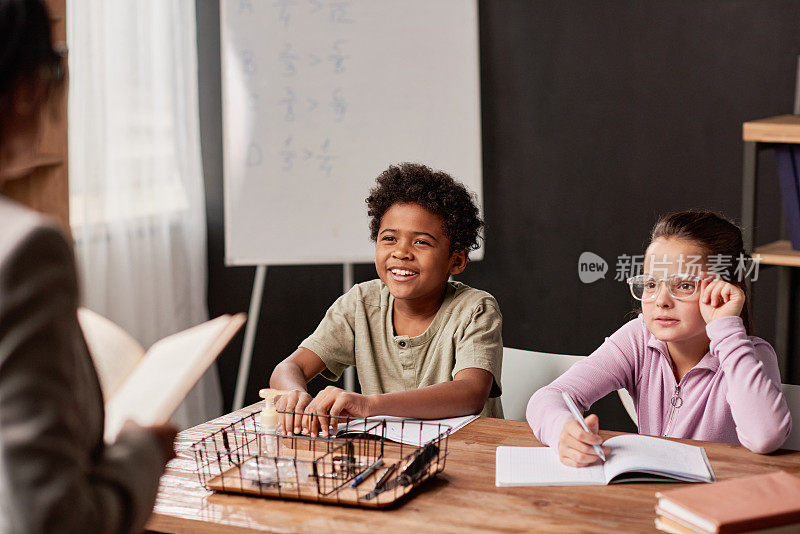 微笑的黑人男孩和严肃的书呆子女孩坐在桌旁，一边听老师讲课，一边拿着练习本