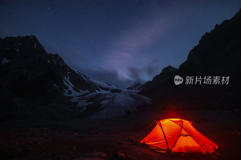 令人敬畏的山地景观与生动的橙色帐篷附近的大冰川舌在夜间星空下的云。帐篷在橘黄色的灯光下闪耀，在星夜中可以看到冰川和山脉的剪影。