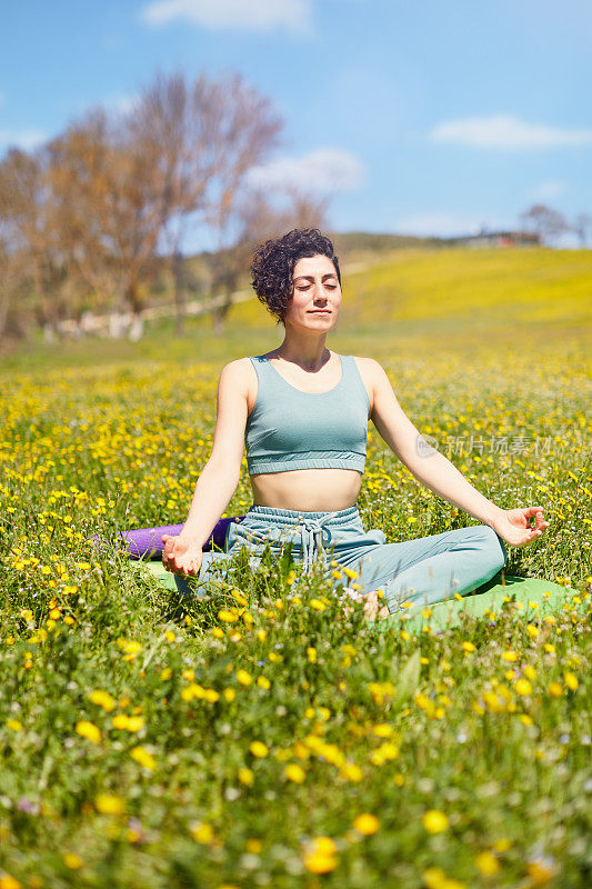 在一个阳光明媚的日子里，一个年轻的女人在满是五颜六色的野花和蓝天的草地上冥想和练习瑜伽。在大自然中寻找灵魂，放松精神