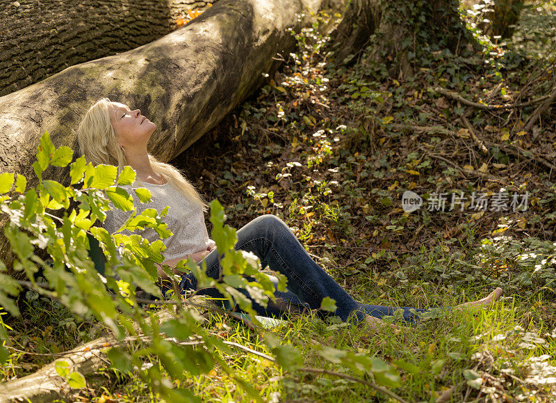 成熟的金发女人放松地倚在阳光下的树干上