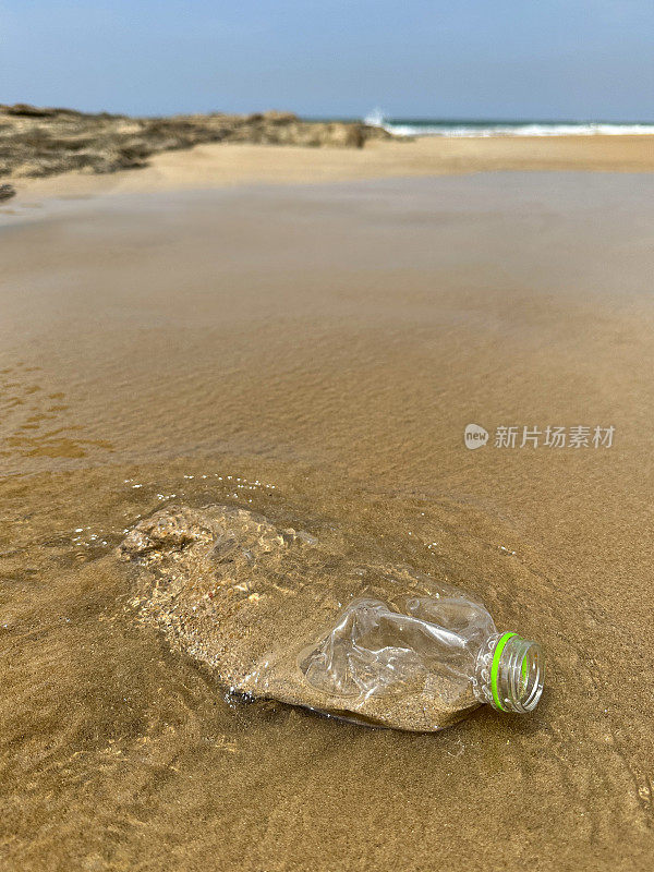 特写图像，粉碎，透明的塑料水瓶没有盖子，含有沙子和水被冲到沙滩上的退潮，海洋垃圾和岸上的污染，乱扔的沙子，肮脏的海滩，关注前景