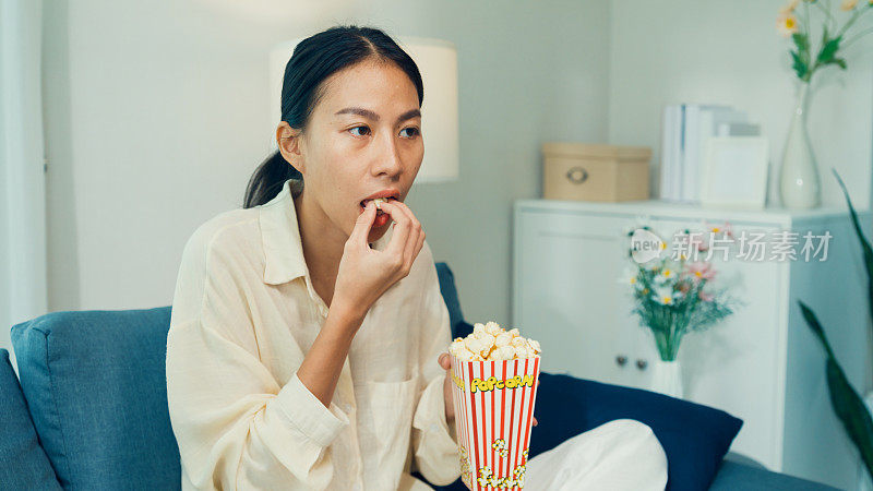年轻的亚洲女孩在沙发上吃爆米花，笑着看喜剧频道的电影或电视上的情景喜剧，在家里在线点播，视频点播。