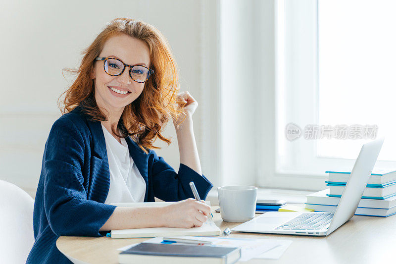 侧影:一头狐色头发的富商女性，微笑着，在记事本上记录信息，喝咖啡，在笔记本电脑上搜索信息，开发创业项目