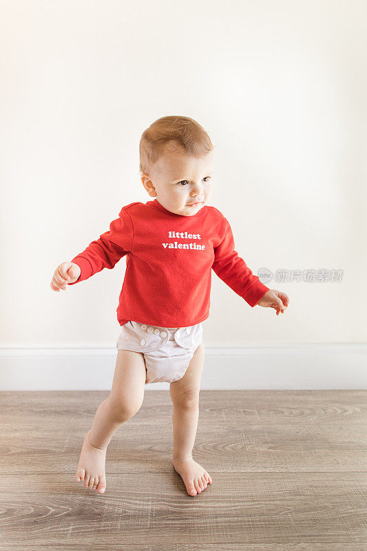 2023年2月的情人节，一个14个月大的可爱男婴赤脚站在木制硬木地板上，穿着一件写着“最小的情人节”的红色毛衣和裸色的布尿布