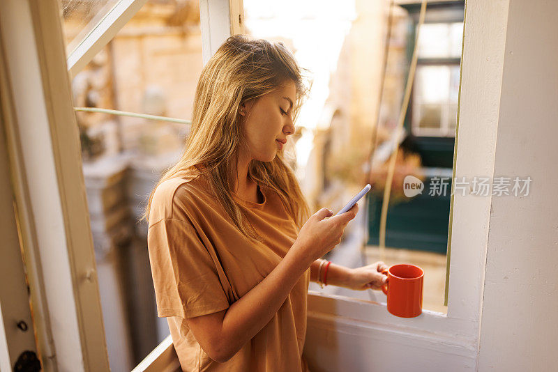 微笑的年轻女子一边在阳台上喝咖啡一边发短信