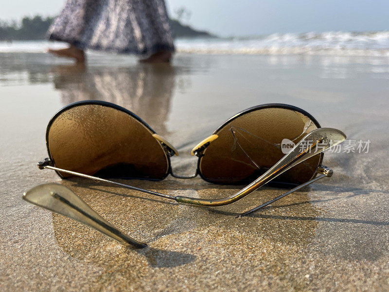 一对飞行员的特写图像，金属框，着色，镜面太阳镜被冲到潮湿的海滩沙滩上，在退潮的水的边缘，破碎的镜头，背景中无法辨认的人，海浪在海岸上破碎，聚焦在前景