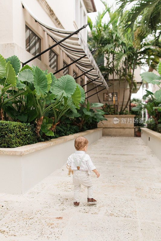 2023年5月，佛罗里达州热带棕榈滩，一个1岁的古巴裔美国小男孩，一头浅棕色短发，戴着鼠尾草绿色领结，穿着背带裤，白色纽扣衬衫，裤子和棕色鞋子，在花园里散步