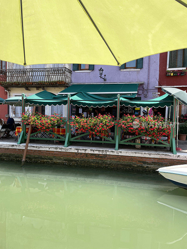 意大利威尼斯布拉诺的威尼斯泻湖岛上的运河图像，露天用餐区装饰着红色的开花植物，系泊的船，重点在前景