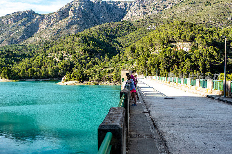 一群游客站在桥上欣赏美丽的风景，蓝色的湖泊和覆盖着森林的山脉。