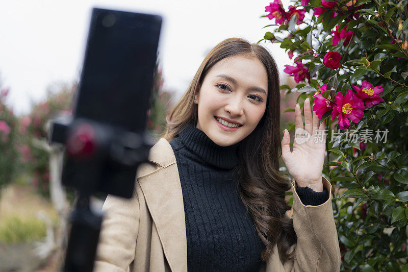韩国一名微笑的旅游博主网红在韩国济州岛的玫瑰花园公园拍摄冬季视频，她正在用智能手机为她的粉丝直播