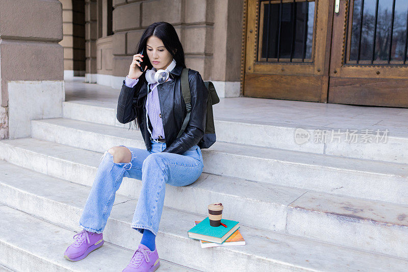 年轻漂亮的女大学生背着双肩包坐在大学楼梯上玩手机。她戴着耳机，穿着皮夹克，等待着大学课程的开始。