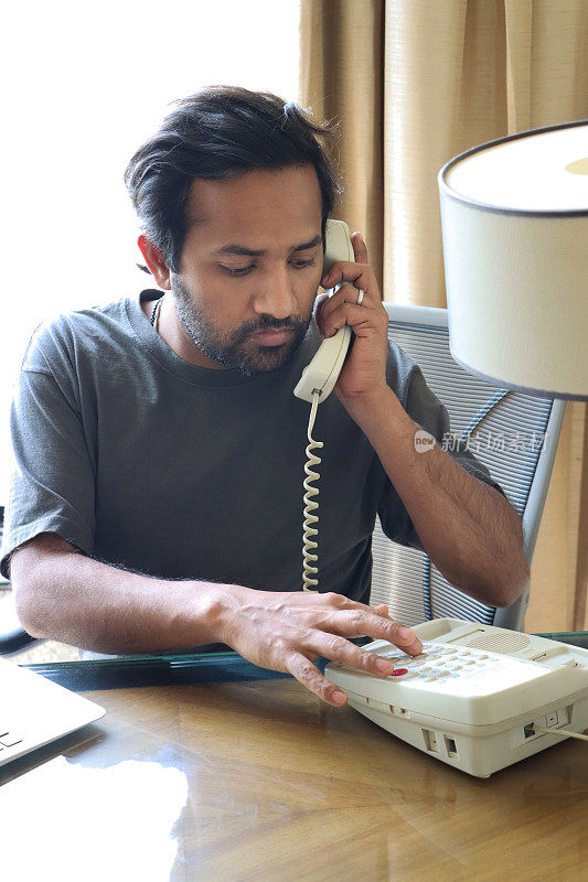 印度人穿着休闲装在办公环境中工作的特写图像，在按键式座机电话上拨号，木制办公桌上的笔记本电脑键盘，办公椅，重点在前景
