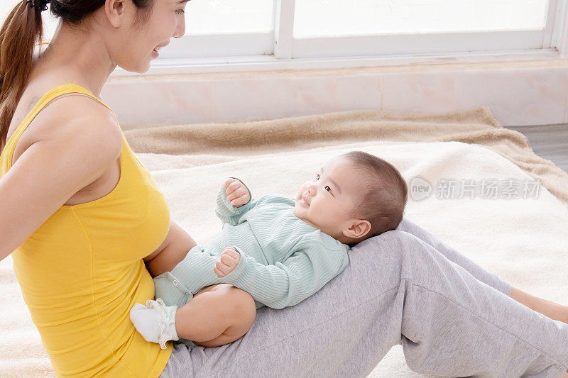 美丽的亚洲妈妈和蹒跚学步的孩子聊天，而孩子躺在妈妈的腿上，年轻的妈妈和她可爱的小宝贝在家里亲密无间。妈妈带着爱和温柔看着婴儿。