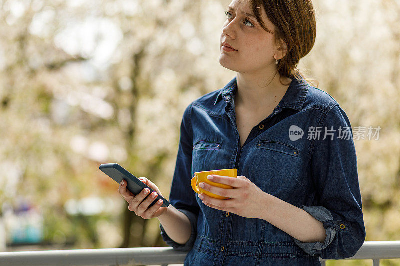 年轻女子站在阳台上，一边喝咖啡一边打电话