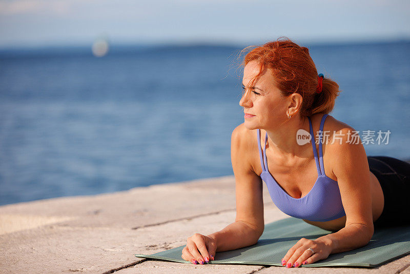 健康成熟的女人躺在海边的垫子上，胳膊肘倚着，望着远方