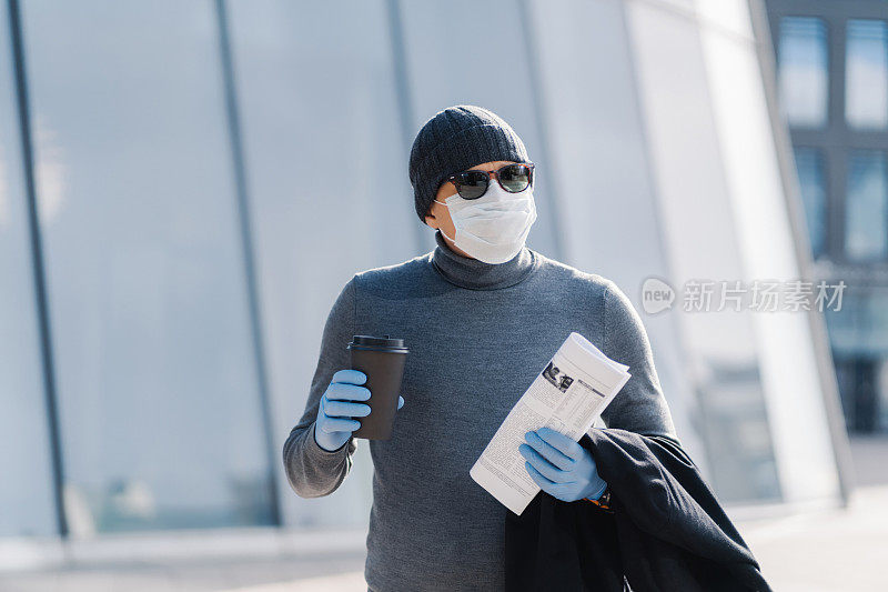 穿着冬装、拿着咖啡和报纸的男子，在现代建筑背景下，展示了戴着手套和口罩的疫情生活