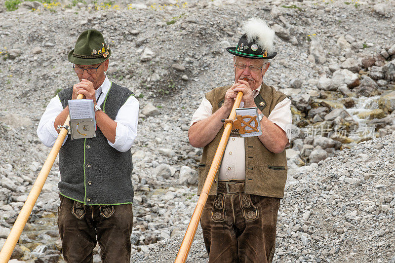 身穿奥地利传统服装的两名男子吹奏圆号。传统的皮裤“皮袜”和蒂罗尔帽。背景中的岩石。阿尔弗恩音乐节。2023年7月8日，奥地利布兰德