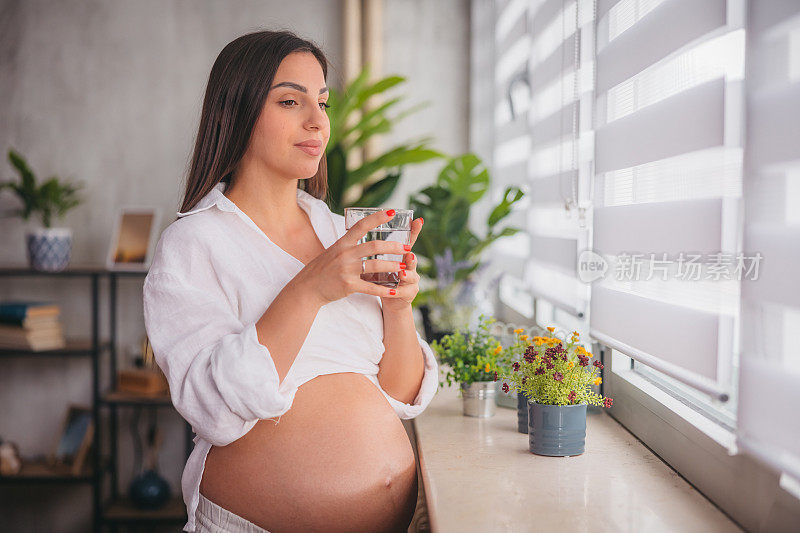 年轻的孕妇一边喝水一边望着窗外