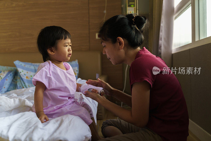 亚洲小男孩用生理盐水点滴坐在病床上，妈妈握着他的手安慰他。可爱的亚洲孩子感染病毒住院了。婴儿保健医疗理念。