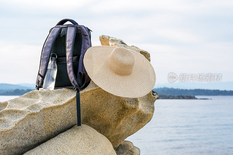 哈尔基季基上,在你的春天徒步旅行。背包，夏季帽和可重复使用的塑料瓶水放在海滩附近的岩石上