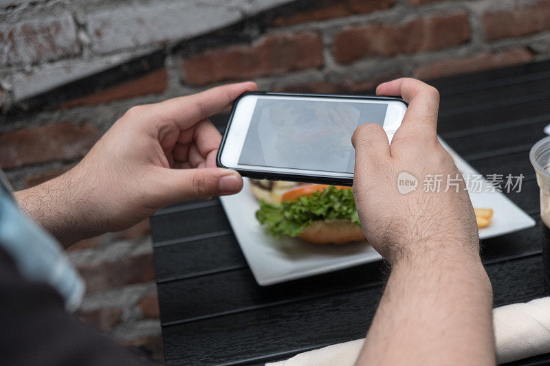 一名男子拿着手机给他的食物拍照。智能手机食品摄影。在一家户外酒吧里给汉堡和炸薯条拍照。