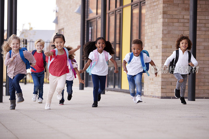 一群微笑的多民族学校的孩子在他们的幼儿学校大楼外的人行道上跑步课后