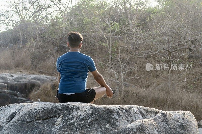 印度男子练习莲花姿势(莲花瑜伽)瑜伽位置在岩石上，后视图，水的边缘，帕洛伦海滩，果阿，印度