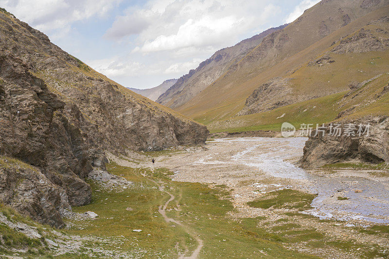 徒步旅行者，一名男子在小河附近的山路上徒步旅行，从靠近中国和吉尔吉斯斯坦边境的山口下到塔什拉巴特