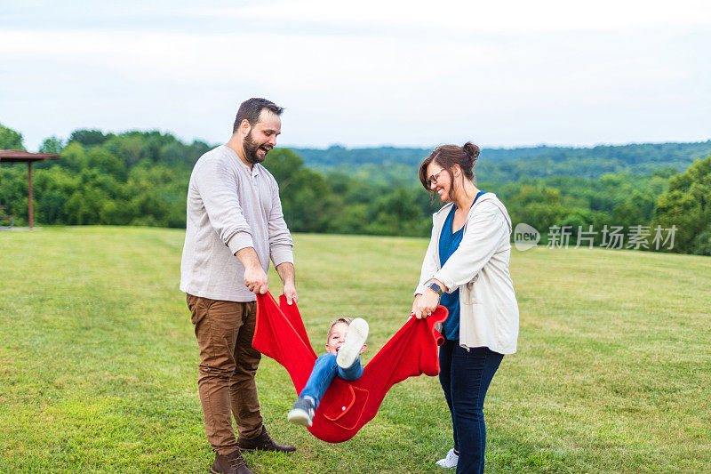 年轻夫妇和儿子玩作为一个家庭在公共草地绿色公园设置一起玩