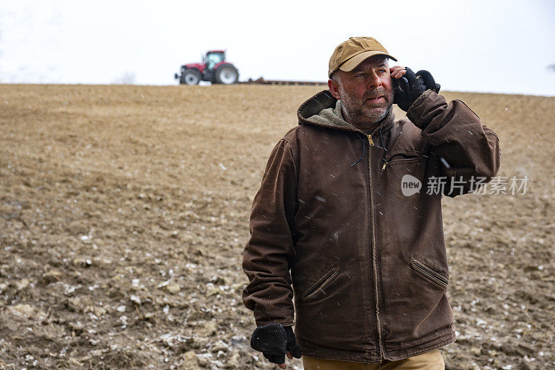 一位成熟的农民站在雨夹雪下的田地里使用手机。