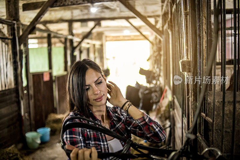 女人准备训练马，站在马厩里准备马鞍-一个年轻女人站在马厩里的马