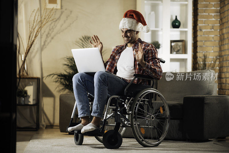 一个年轻人坐在轮椅上与圣诞老人的帽子在一个视频电话祝愿圣诞股票照片快乐
