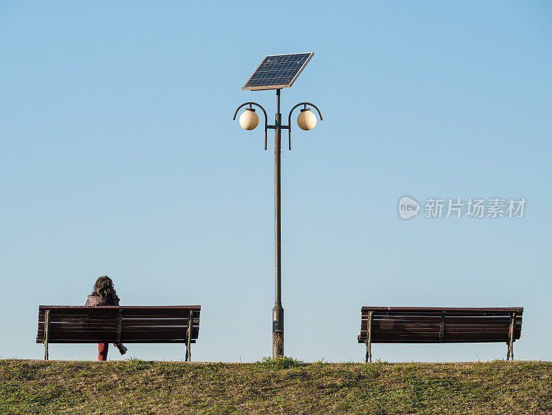一个孤独的女人坐在电线杆旁的长椅上。两张长凳中间有一个太阳能灯柱