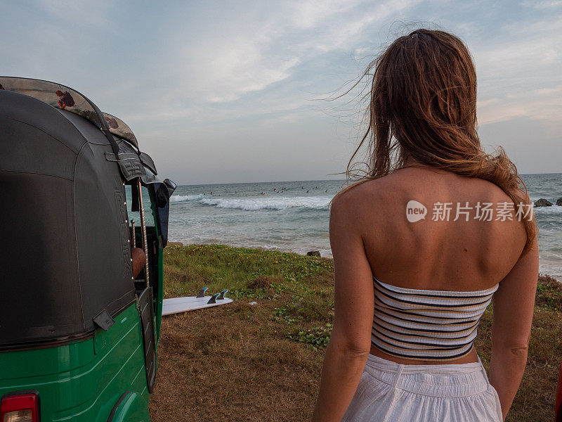 一名妇女骑着人力车停在当地的海滩上，观看远处的冲浪者