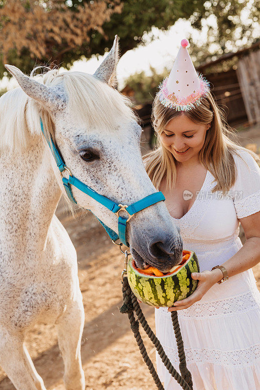 年轻的金发波西米亚女孩穿着白色波西米亚连衣裙，戴着派对帽庆祝她自己的生日和她的马的生日，喂它一个装满水果和蔬菜的西瓜